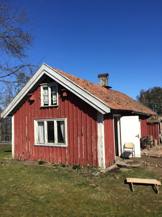 Ett gammalt tvåvånings torp med rödfärgad fasad och tegeltak som behöver renoveras, omgivet av grönska och en klarblå himmel.