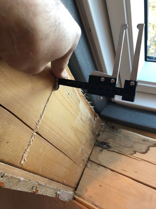 Hand mäter djupet på sågat jack i träbjälke nära takfönster med skjutmått.