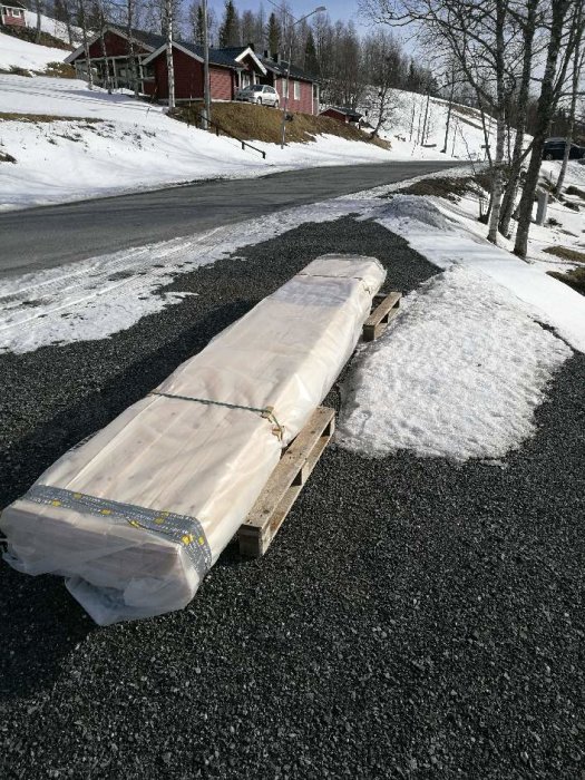 Inplastad limträbalk på en pall vid en snöig vägkant med röda hus i bakgrunden.