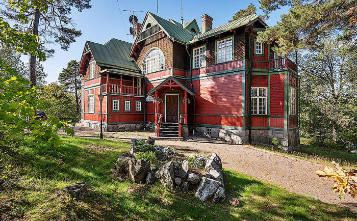 Rött och grönt trähus med jugenddetaljer i soligt Djursholm, omgivet av träd och stenmur på framsidan.