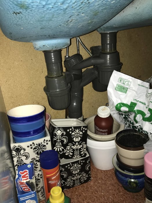 Gamla vattenlås under diskbänk med diverse hushållsartiklar och förpackningar runtomkring.
