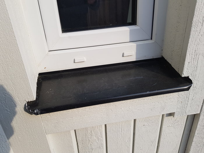 Ett svart fönsterbleck under ett vitt fönster med korrekt vattenavrinning, installerat in under sidpanelerna på en husvägg.