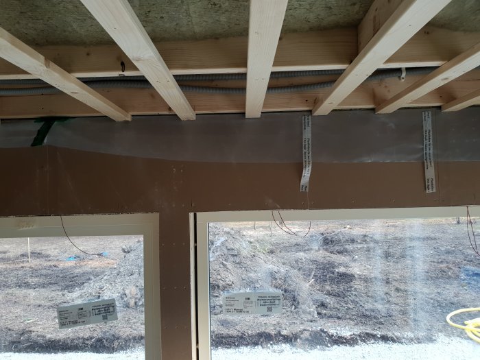 Inomhus byggkonstruktion med exponerade träbjälkar, oöppnad diffusionsspärr och utsikt genom fönster.