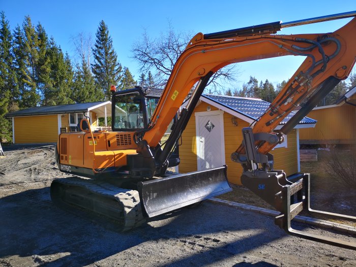 Orange grävare på en tomt med skopa fram och skog i bakgrunden.