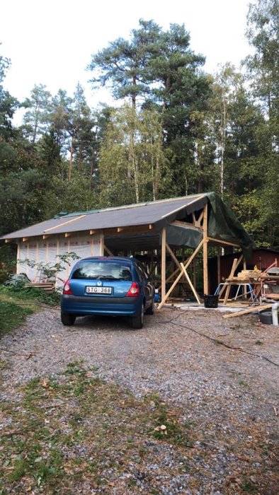 Halvfärdigt dubbelgarage med en bil framför, i en skogsmiljö, den vänstra delen med tak och väggar, den högra under konstruktion.