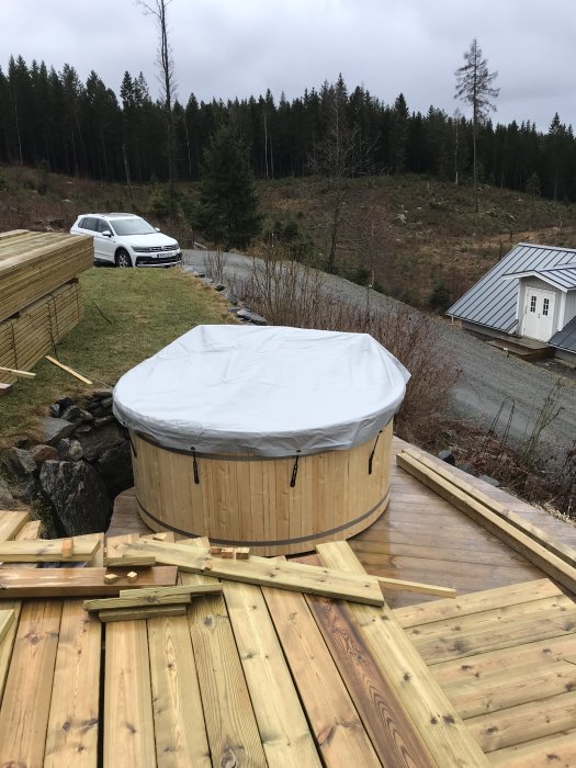 En tomt med lutning visar en badtunna, en trätrall framför och en vit bil parkerad på grusvägen nedanför.