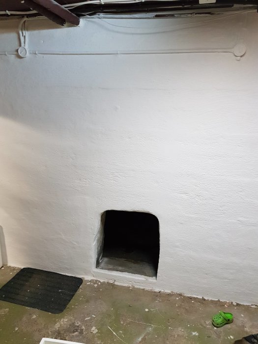 Vit källarvägg med ett litet kvadratiskt hål som leder in under verandan.