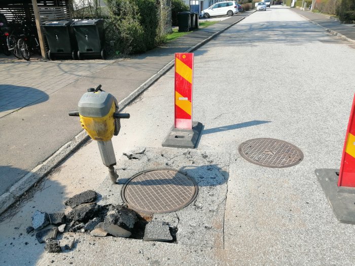 Gatuborr står nära uppbruten asfalt och brunnslock med varningsskyltar vid arbetsområde.