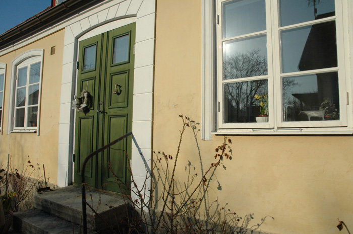 Ytterdel av gult hus med grön dörr, fönster och vit omfattning under solig himmel.