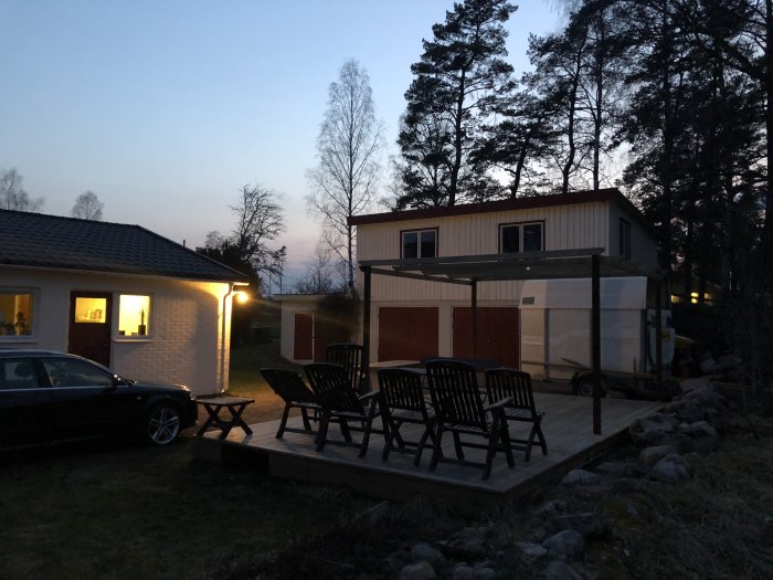 Kvällsbild av en trädäck uteplats med trädgårdsmöbler framför ett hus, med grannhus i bakgrunden.