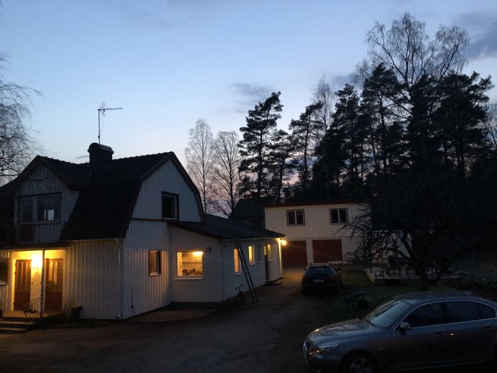 Kvällsbild av ett bostadshus och en garagebyggnad med några tända lampor och en bil parkerad framför huset.