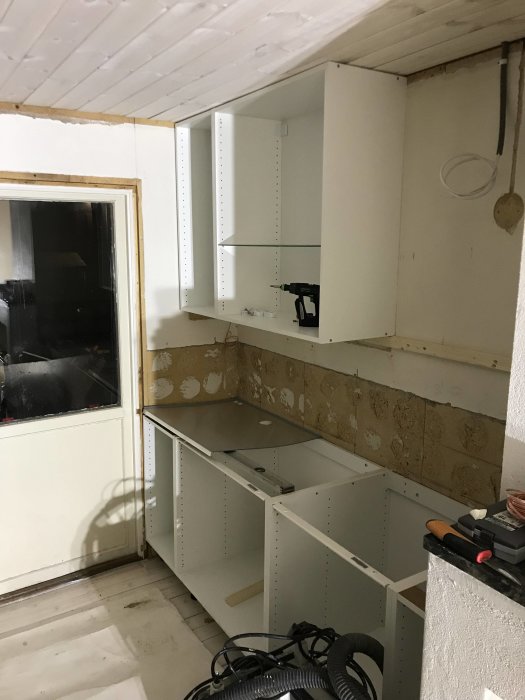 Halvfärdigt köksstomme i en renovering, vita skåpsstommar med en glashylla och verktyg på golvet.