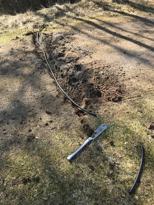 Reparerad gräskant med synlig svart kabel, ett öppet rör och en spade på en grusväg.