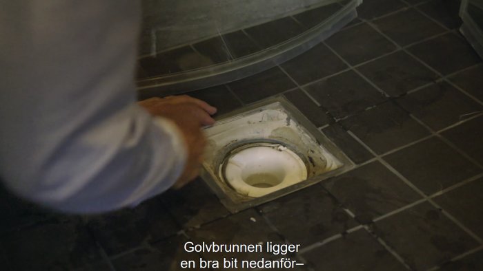 Person som pekar på en golvbrunn inbyggd i ett klinkergolv, vilket illustrerar diskussionen om korrekt VVS-installation.