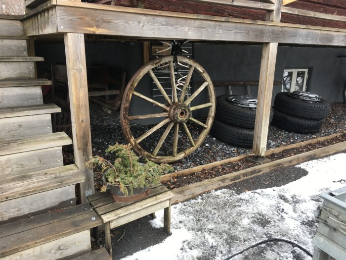 Två upphängda sommardäck under en trätrappa med snöfläckar på marken och ett gammalt vagnshjul.