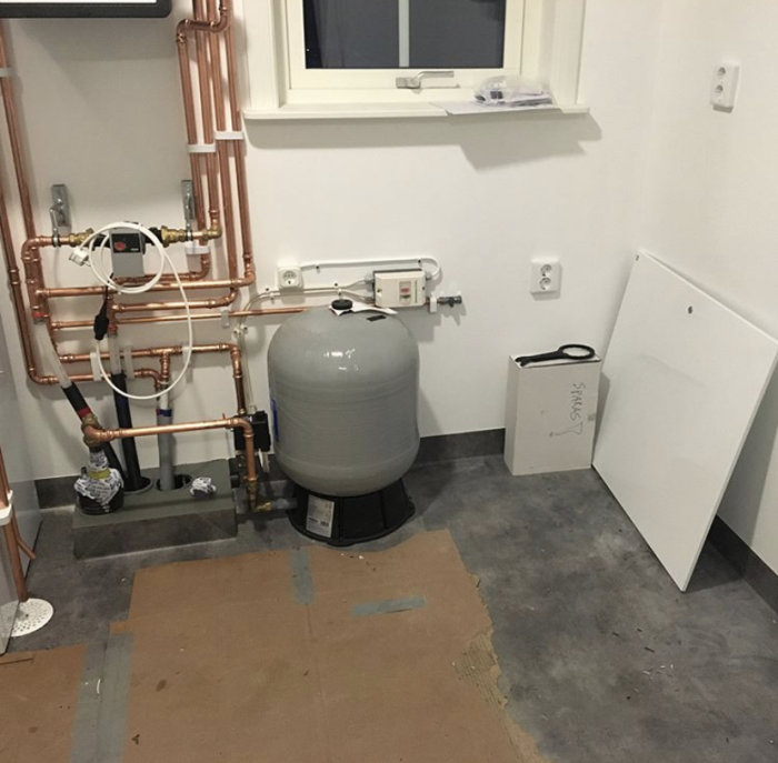 Bergvärmepump och varmvattenberedare installerade i ett hörn av en tvättstuga, omgivna av kopparledningar och skyddade med ett öppningsbart skåp.