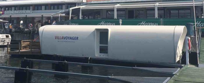Hus på en flotte vid brygga med texten "VILLAVOYAGER" och en bakgrund med båtar och kaj.