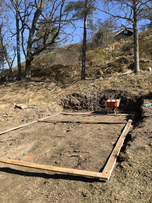 Förberedd mark för bygge av attefallshus med synliga träramar på en sluttande tomt med träd och ett hus i bakgrunden.