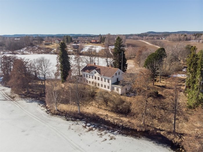 Flygbild på tvåvåningshus vid frusen å omgivet av träd och snötäckt landskap.