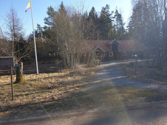 Röd lada med grusad gårdsplan omgiven av träd och en flaggstång med prideflagga.