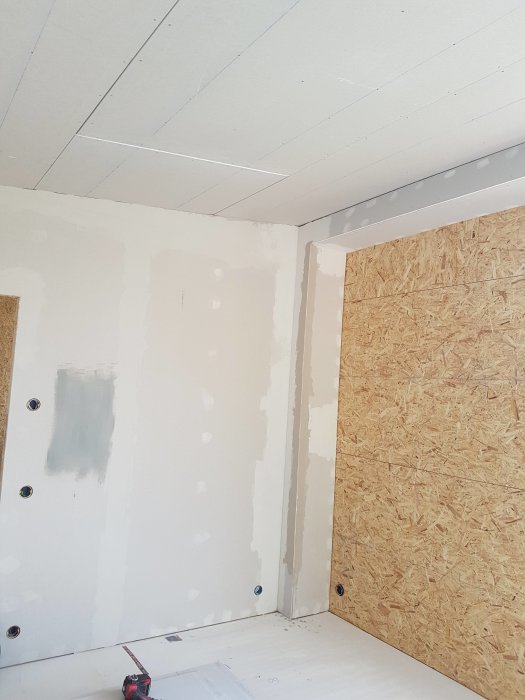 Hörn av ett rum under renovering med oslipat spackel på väggarna, OSB-panel och uppsatta gipsplattor i taket.