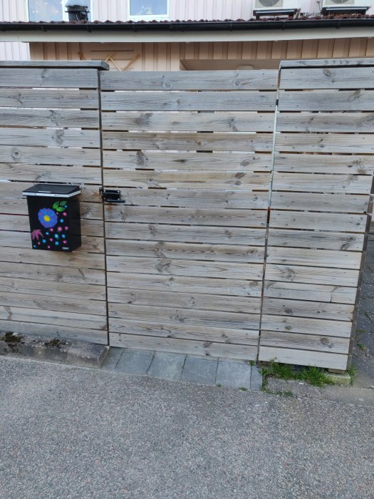 Trästaket med oregelbundna plankor och en dekorerad soptunna vid en stenlagd trottoar.