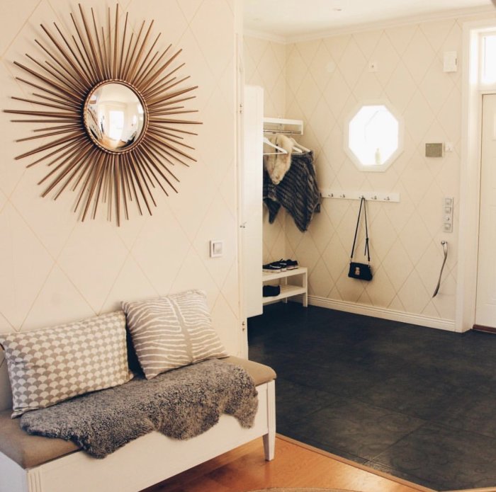 Elegant hall med spegel på vägg, mönstrad tapet, bänk med kuddar och entrémöbler.