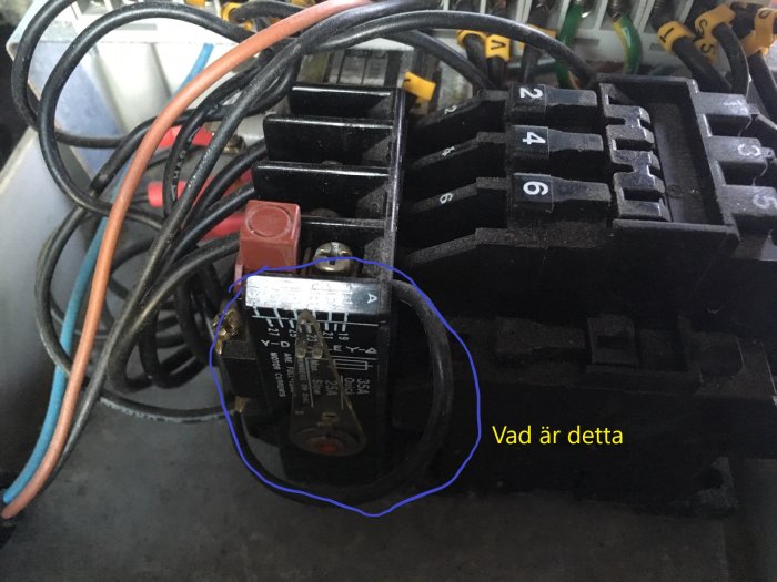 Närbild av en komponent markerad med blått på en elektrisk pump, omgivet av kablar och anslutningar, med texten "Vad är detta".