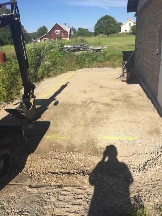 Grävmaskin nära utgrävd mark vid en baksida av ett garage med märkfärg och skugga av personen som fotograferar.