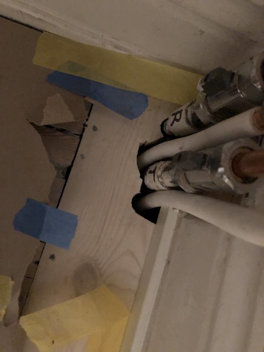 Vägg med överdimensionerade hål runt rörledningar med synlig isolering och blått samt gult tejp.