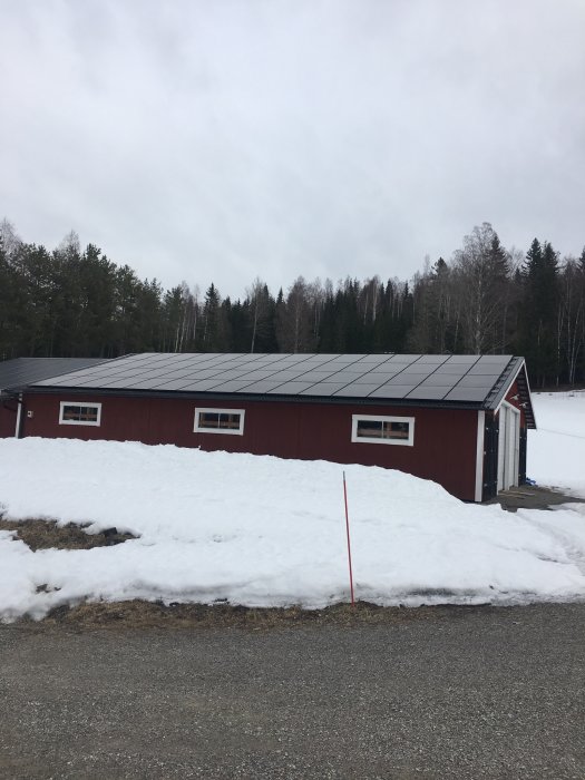 Röd träbyggnad med solpaneler på taket omgiven av smältande snö och träd i bakgrunden.