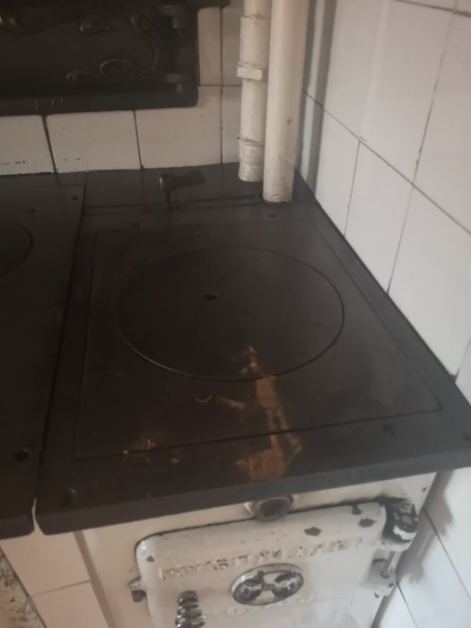 Gammal spis med reglage och stängt lock på ovannämnd stående vedeldad köksspis, bredvid vit kaklad vägg.