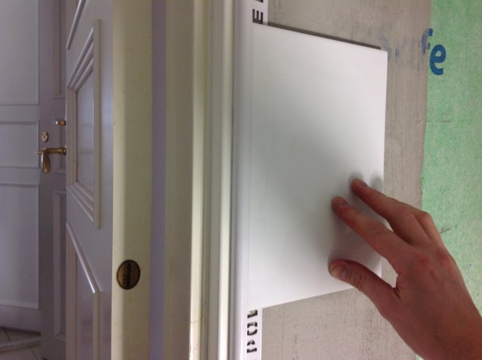 En persons hand rör vid vit kakelplatta nära en dörr med mörk handtag och namnskylt.