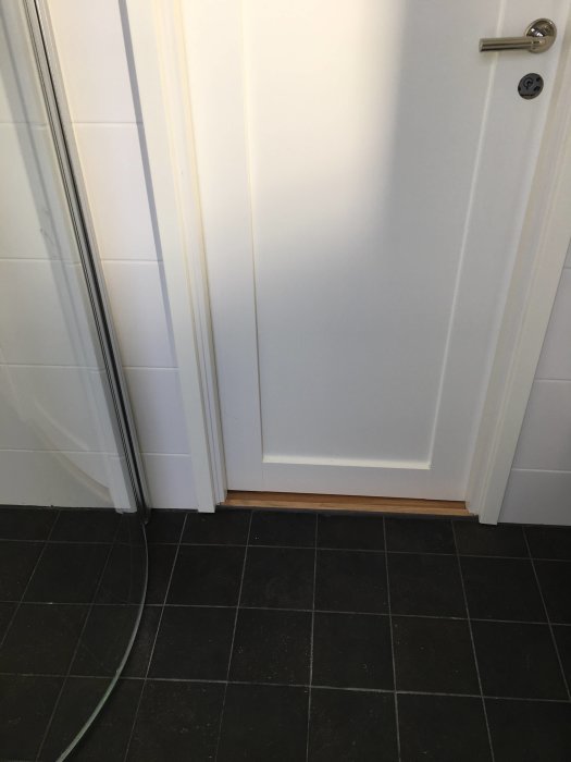 Plastklackfoder vid dörrtröskel i våtutrymme med vita väggar och mörkt klinkergolv.