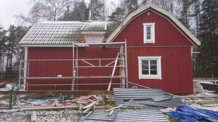 Rödt timmerhus under renovering med byggnadsställning och gammal takplåt spridd på marken.