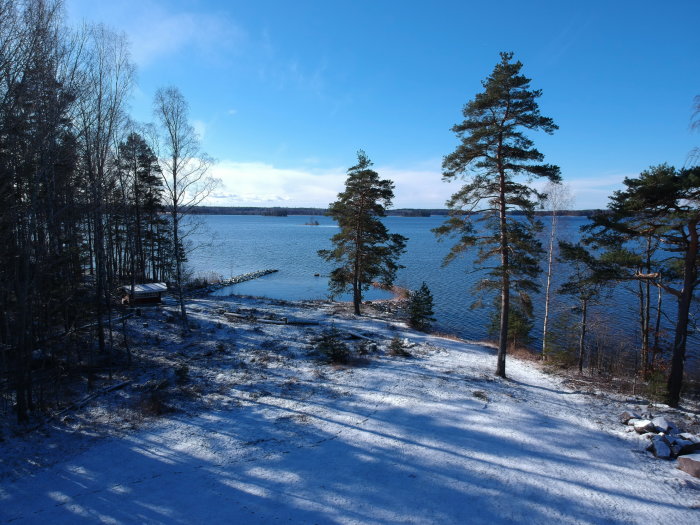 Vinterutsikt över en strandtomt med snö, träd, en brygga och en klarblå sjö under en solig himmel.