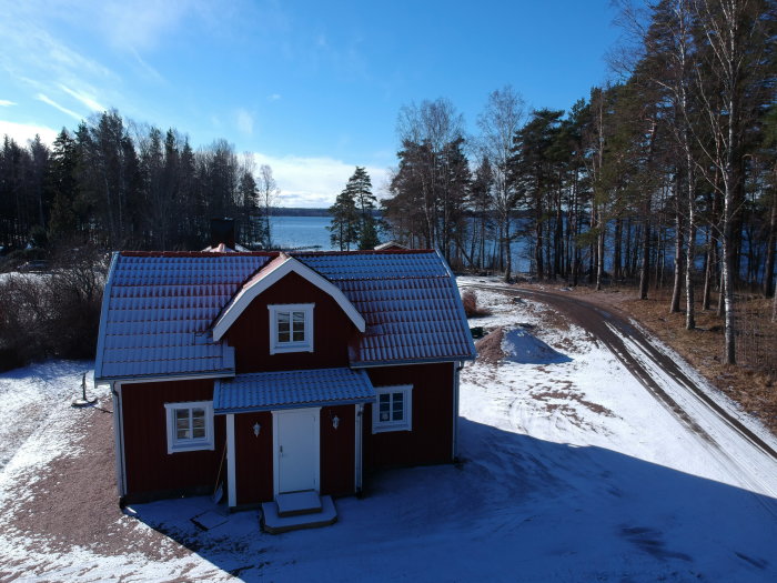 Rödfärgat hus med snötäckt tak nära en strand och en skogsväg i vintersol.