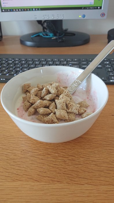 En skål med fullkornsflingor och yoghurt på ett träbord med en dator i bakgrunden.