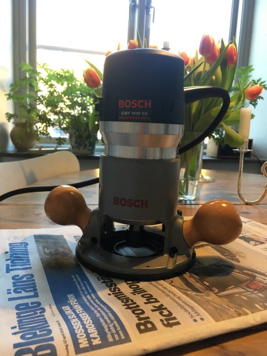 Bosch GMF 1400 CE fräsmaskin på ett bord framför fönster med tulpaner och krukväxter.