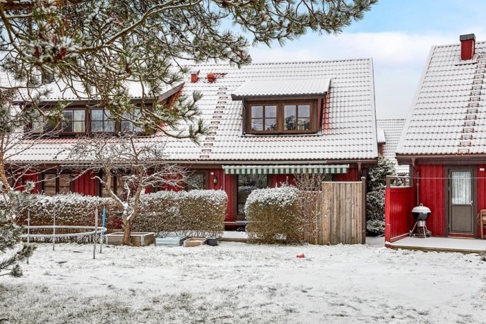 Rött 70-talshus med snötäckt tak och trädgård under vintern, tydligt i ett villaområde.