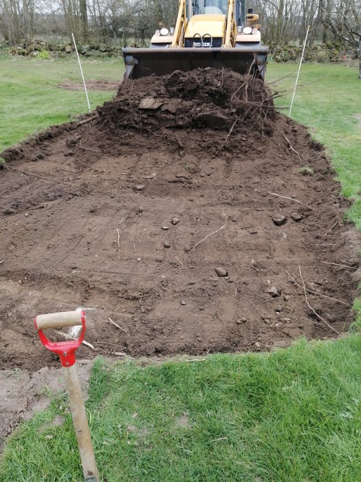 Pågående arbete med att gräva ut marken för ett växthus med en grävmaskin i bakgrunden och en spade i förgrunden.