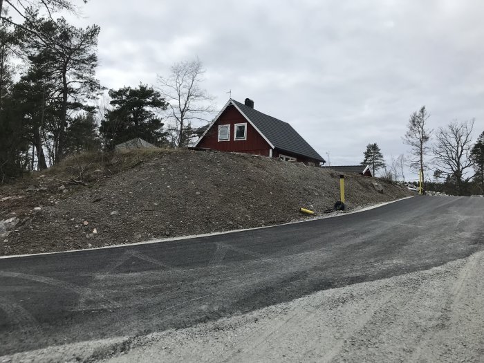Slänt med grus och sparsam vegetation vid en nyasfalterad väg, röd stuga i bakgrunden.