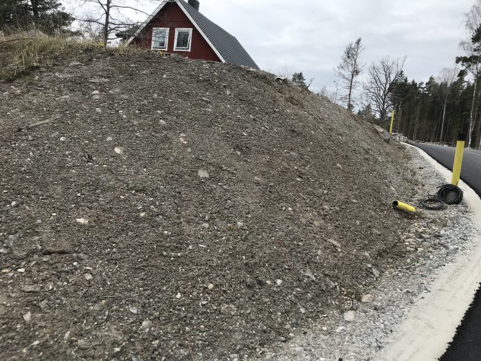 Ojämn slänt av grus och stenar framför röd stuga efter vägarbete, i behov av landskapsförslag.