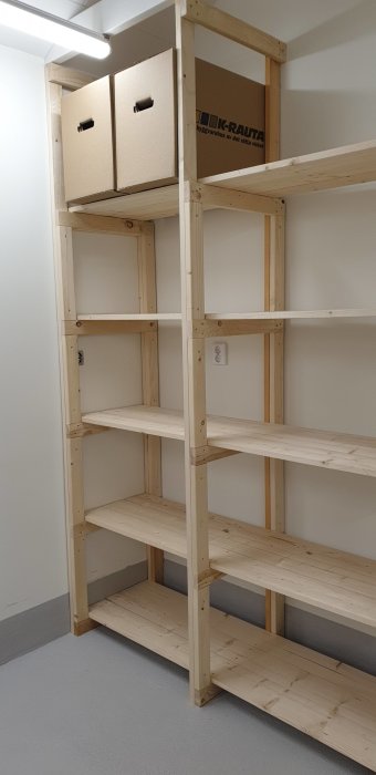 Hyllor av trä i ett förråd med två kartonger på översta hyllplanet.