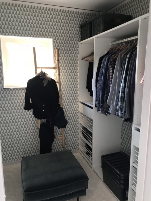 Walk in closet med välorganiserade klädhängare, en svart sittpuff och geometriskt tapetmönster.