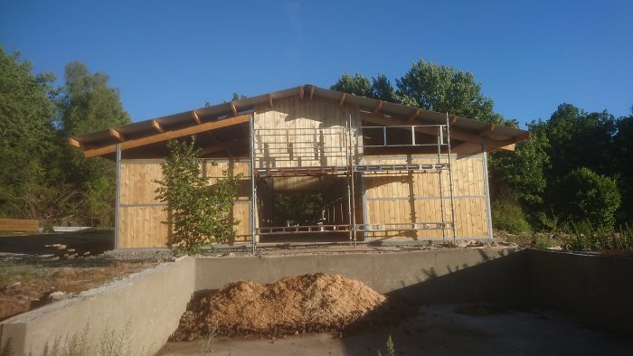 Stall under konstruktion med takplåtar, bärlina, klädselbräder och ställningar under en klar himmel.