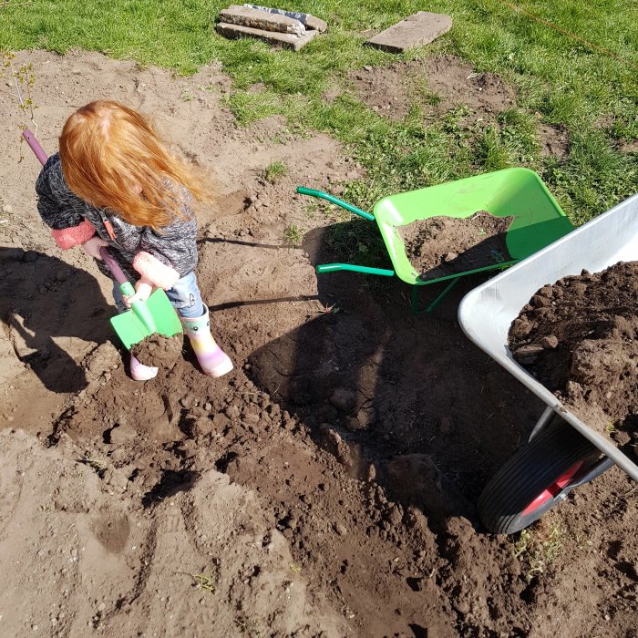 Barn som gräver i en rabatt med en grön spade, bredvid en grön skottkärra fylld med jord.