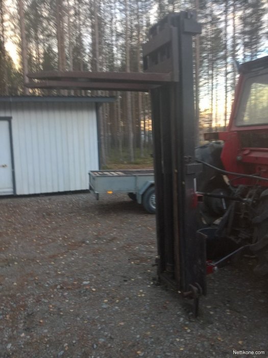 Hemgjord lyftanordning med trucktorn monterad på en liten traktor framför en släpvagn och förrådsbyggnad.