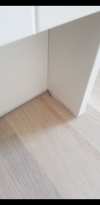 Dåligt applicerad genomskinlig silikon längst in vid skåpets och väggens möte ovanför ett trägolv.