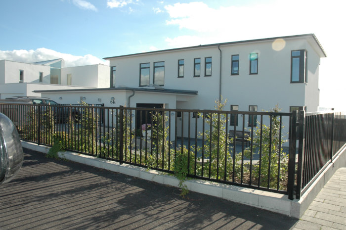 Svart metallstaket med vertikala spjälor runt en trädgård framför modern vit husfasad.